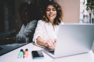 Beyaz ceketli, netbook 'u karıştıran, modern binanın cam duvarının yanındaki kafede oturmuş gülümseyen şık genç bir kadın.
