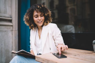 Cep telefonuyla mesajlaşırken ve caddedeki kafedeki ahşap masada otururken kıpır kıpır kahverengi saçlı, gülümseyen ve bakışlarını çeviren olumlu bir genç kadın.