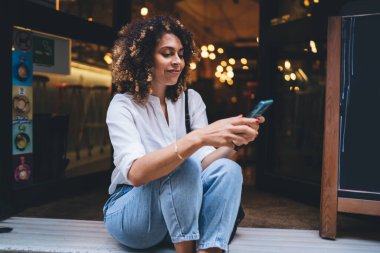Neşeli, pozitif, günlük kıyafetler giyen, haftasonu kafede dinlenirken yerde oturup akıllı telefonlara bakan bir kadın.