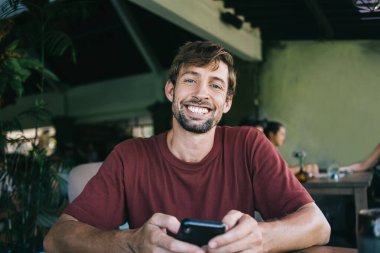 Kafkasyalı neşeli bir hippinin yarım boy portresi sohbet etmek ve blog yazmak için cep telefonu kullanıyor, elinde cep telefonuyla kameraya bakıp mesajlar yolluyor.