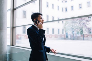 Düşünceli genç etnik işadamının cep telefonuyla konuşması ve modern ofiste büyük cam pencerenin yanında durması.