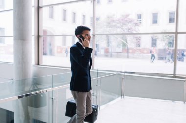 Ciddi etnik iş adamlarının resmi giysileri ve gözlükleriyle modern ofiste bavulla durup akıllı telefondan konuşmalarının yan görüntüsü