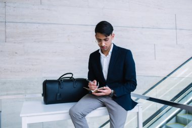 Resmi evrak çantası ve gözlüklü düşünceli etnik erkek girişimci yürüyen merdivenin yanındaki beyaz masada oturuyor ve notlar yazıyor.