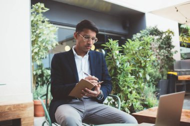Resmi takım elbiseli ve gözlüklü genç adam modern binanın terasındaki yeşil bitkilerin yanındaki sandalyede oturuyor ve deftere yazı yazıyor.