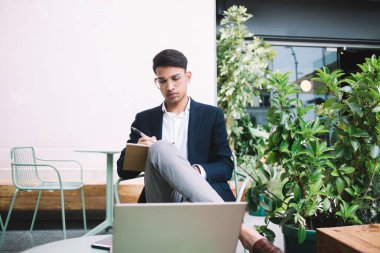 Resmi takım elbiseli ciddi bir etnik adam bacak bacak üstüne atmış ve balkonda dizüstü bilgisayar projesinde çalışırken deftere not alıyor.