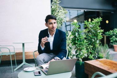 Resmi takım elbiseli mutlu Asyalı genç adam modern kafedeki terasta kameraya bakarken dizüstü bilgisayar ve belgelerle masada oturuyor.