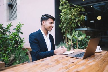 Resmi takım elbiseli, ahşap masada dizüstü bilgisayarı ve bir fincan taze kahveyle oturan ve planlamacıda notlar alan mutlu etnik genç adam, dışarıda modern kafenin koyu cam duvarına ve saksı bitkilerine karşı çalışıyor.