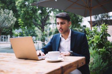 Modern kendine güvenen yakışıklı iş adamı yaz tatilinde öğle yemeği molasında açık kafede dinlenirken elinde lezzetli bir fincan kahveyle dizüstü bilgisayarla sörf yapıyor.