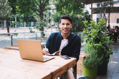 Neşeli gülümseyen genç bir erkek öğrenci, günlük kıyafetlerini giyer, yazı defterinde notlar yazar ve gündüz vakti rahat bir kafedeki yeşil bitkilerin arka planında dizüstü bilgisayarı ve cep telefonuyla ahşap masaya oturur ve kameraya bakar.