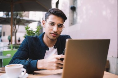 Yakışıklı etnik erkek serbest, şık saçlı, akıllı telefon kullanan ve gündüz kafede dizüstü bilgisayarla çalışırken kahve içen.