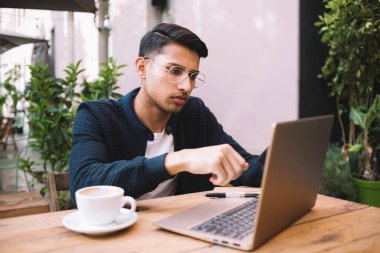 Sıradan giyinen, gözlüklü, dizüstü bilgisayarda gezen ve kafede kahvenin keyfini çıkarırken kalem kullanan konsantre genç bir etnik adam.