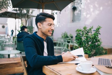 Gülümseyen, gözlüklü ve günlük giysileri olan genç bir erkek sokak kafesinde oturuyor ve uzaktan çalışırken cep telefonuna bakıyor.