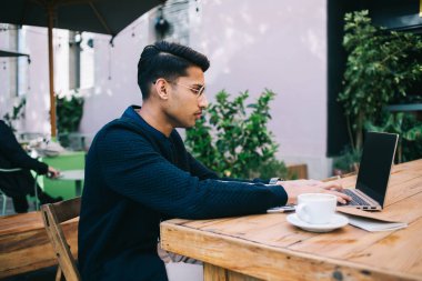 Gözlüklü ve gündelik kıyafetlerle dizüstü bilgisayarda gezinen, uzaktan proje üzerinde çalışan ve sokak kafesinde oturan ciddi genç bir erkeğin yan görüntüsü.