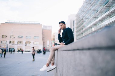 Kendine güveni tam bir erkek, şık giyiniyor ve gözlük takıyor. Şehir sokağındaki modern binanın yanında oturuyor. Klasik evlerle ve telefonda konuşurken kameraya bakıyor.