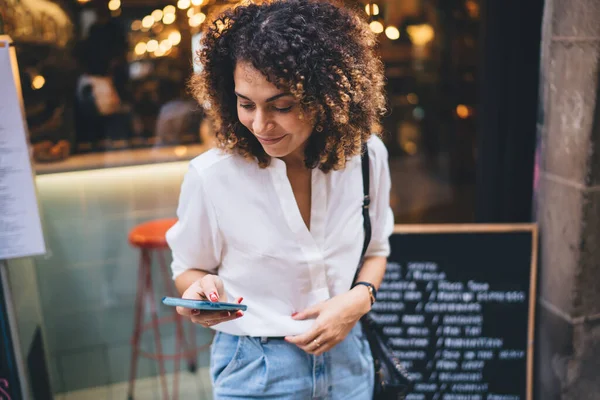 スマートフォンの笑顔と画面を見てカジュアルな服を着てカフェの近くの通りに立っている間にインターネットを閲覧すると肯定的な民族の女性 — ストック写真