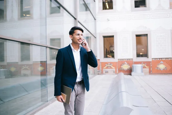 Cheerful hindu male entrepreneur in trendy wear and outfit making mobile phone call in roaming connection outdoors, smiling businessman dressed in stylish wear and spectacles talking on cellular