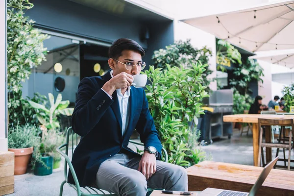 Serious prosperous male entrepreneur in trendy wear looking at camera drinking aroma coffee on remote job, pensive confident businessman sitting on cafe terrace doing freelance jon use laptop