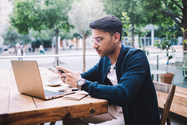 Вид сбоку на молодого этнического мужчины в повседневной одежде и очках с помощью смартфона во время работы дистанционно с ноутбуком в уличном кафе днем