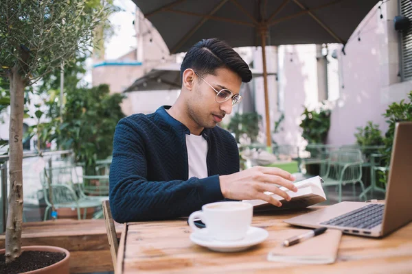 穿着休闲装和眼镜的年轻男性聚精会神地坐在街边咖啡馆的桌旁看书 同时用笔记本电脑学习 — 图库照片