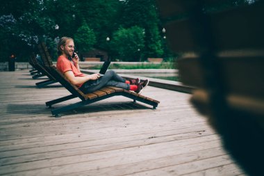 Günlük kıyafetli neşeli bir adamın yan görüntüsü dizüstü bilgisayarlı, dinleniyor ve parktaki rıhtımdaki telefona cevap veriyor.
