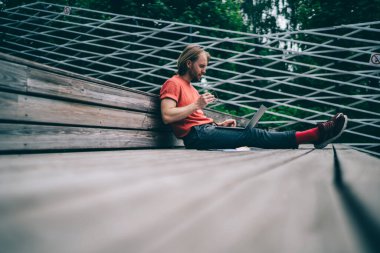 Genç adam dizüstü bilgisayar ekranına uzanmış bacaklarıyla bakıyor ve tahta zeminde otururken elinde gözlük tutuyor.