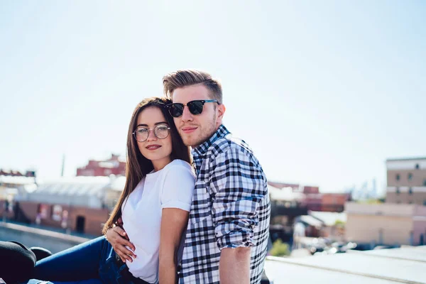 Portrait of Caucasian couple in love in trendy spectacles and sunglasses looking at camera during date cuddling, casual dressed hipster guys 20 years old posing during together pastime at house roof