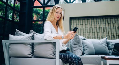 Sosyal medyada cep telefonuyla mesajlaşan ve masanın yanında dizüstü bilgisayarla oturup kameraya bakarken internette sörf yapan mutlu bir kadın.