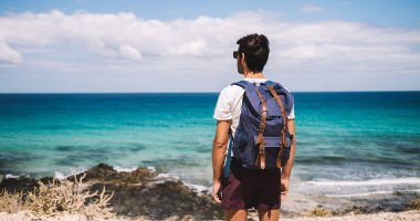 Sahilde sırt çantasıyla gezen ve ufka bakarken manzaralı deniz manzaralı gezgin bir erkek.