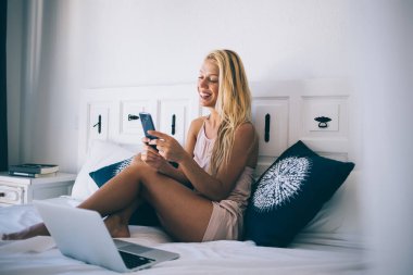 Neşeli kadın serbest çalışan cep telefonu cihazından gelen e-postayı kontrol eden uzaktan çalışan, mutlu bir kadının internet sohbeti sırasında cep telefonu mesajları göndermesi için bilgisayarla rahat bir yatakta oturuyor.