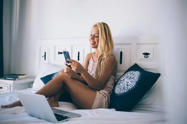 快乐的女性自由职业者在积极的网络聊天过程中 与笔记本电脑坐在舒适的床上 快乐的女性在积极的网络聊天过程中发送手机短信 — 图库照片
