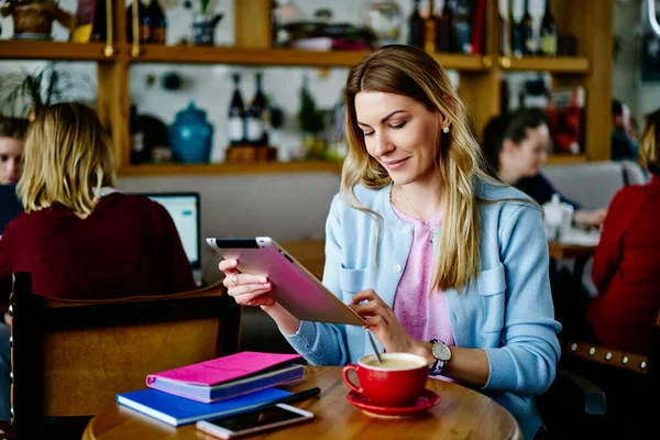 穿着休闲装的积极女性一边微笑着浏览着平板电脑 一边坐在木制餐桌旁喝咖啡和在咖啡店工作 — 图库照片