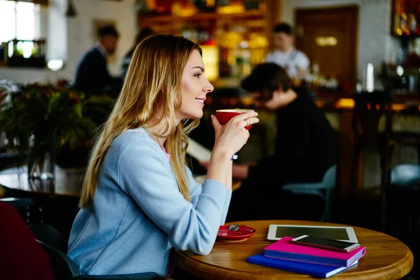 カジュアルな服装で幸せな女性の側面ビューホットコーヒーのカップと木製のテーブルに座って離れて現代のカフェテリアで見て — ストック写真