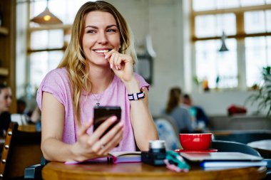 Gündelik giyinmiş genç bir kadın telefon kullanıyor ve bir fincan sıcak içecekle kafede oturuyor ve gözlerini kaçırıyor.