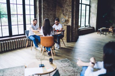 Geniş ofisteki iş fikirleri hakkında konuşurken yuvarlak masada oturan ve çalışan odaklı çok ırklı iş arkadaşlarının tam uzunluğu