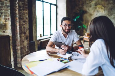 Gündelik kıyafetler ve gözlüklerle ahşap masada oturup modern çatı katında projeleri tartışan neşeli meslektaşlar.