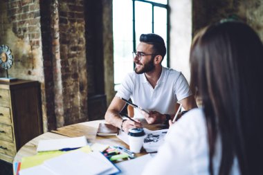 Beyaz tişörtlü ve modern gözlüklü genç şık sakallı adam iş arkadaşıyla kafede oturuyor ve not alırken tablet kullanıyor.