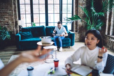 Sıradan giyinen erkek, tabletle kanepede otururken, kadın, modern çalışma alanındaki kağıtlarla masadaki meslektaşıyla konuşuyor.