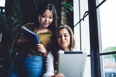 Pencere kenarında oturan, tablet kullanan, yanında not defteriyle tablete bakan Asyalı kadın bir kadının alçak açılı görüntüsü.