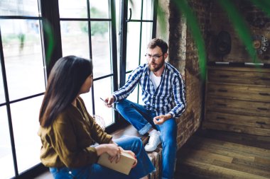 Normal giyinen, gözlüklü, pencerenin kenarına yaslanmış ciddi bir adamın yüksek açılı görüntüsü ve ofisteki yeni başlayan fikirlerle ilgili kadın iş arkadaşıyla konuşması.