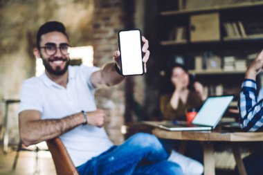 Gözlüklü neşeli adam rahat bir kafede iş arkadaşlarıyla masaya oturup kameraya gülümserken kameraya telefon ekranını gösteriyor.