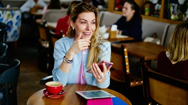 From above of positive young woman in casual clothes smiling and looking away while having video conversation via smartphone during remote work in cozy cafe