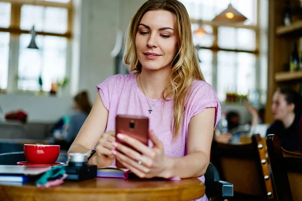 Sarı saçlı genç bir kadın SMS 'e cevap vermek için telefon kullanıyor ve kafede çay içiyor.