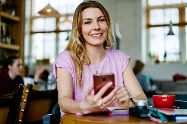 Günlük giyinen genç bir kadın mesaj yazmak için telefon kullanıyor ve kafeteryada dinlenirken bir fincan sıcak içecekle masaya oturuyor ve kameraya bakıyor.