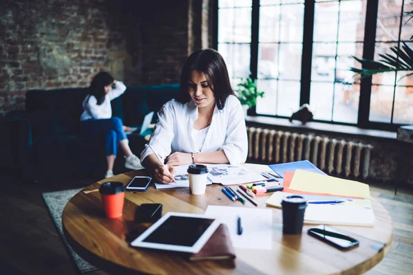 Concentrated female employee in casual clothes sitting at wooden table and taking notes in copybook while working in creative office