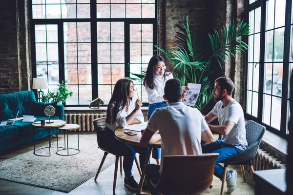 Happy Asian businesswoman in casual wear standing at wooden table and showing document to multiracial coworkers in modern loft office