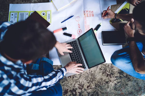 从上面看穿着休闲装的匿名男性创业者坐在地板上 与同事和周围的报纸一起工作在办公室的笔记本电脑上 — 图库照片