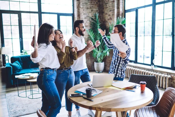 勝利を祝いながら腕を上げてオフィスのテーブルの周りに笑顔と立ってカジュアルな服装で幸せな多人種の同僚のグループ — ストック写真