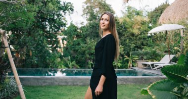 Siyah elbiseli genç, uzun saçlı bir kadın. Yüzme havuzlu, sessiz bir tropikal bahçede kameraya bakıyor.