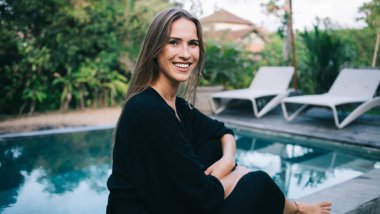 Yüzme havuzunun yanındaki taştan sınırda oturan, siyah takım elbiseli, ince ayaklı, çıplak kadın kameraya bakıyor.