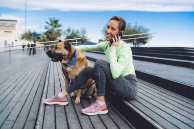 Aktif kıyafetli neşeli bir kadın gülümsüyor ve köpeğiyle birlikte güneşli bir günde sokakta oturuyor ve cep telefonuyla konuşuyor.